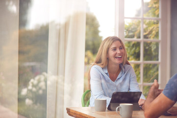 Couple With Digital Tablet Sitting At Table Working From Home Viewd Through Window