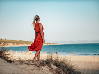 Pretty Blond Girl wearing a long red dress on the beach, playing with sand and looking towards the...