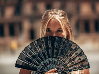 Blond Girl standing in Sevilla holding a Fan in front of her face Showing her Blond hair and  Green brown Eyes