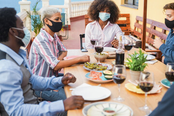 Young multiracial people eating and drinking together while wearing face protective masks - Focus...