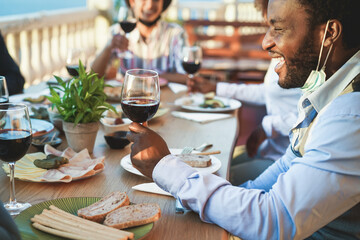 Cheerful multiracial friends eating and drinking wine while wearing protective masks during coronavirus outbreak - Social distance concept - Focus on black man