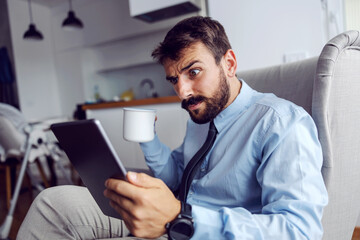 Young shocked father in suit sitting at home in chair, drinking coffee and reading mails.