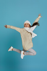 Excited smiling cheerful little child girl in white winter knitted hat and sweater jumping on blue...