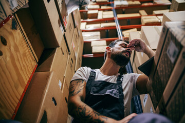 Tattooed bearded worker in overalls having hard day on the work. Storage of import and export firm...