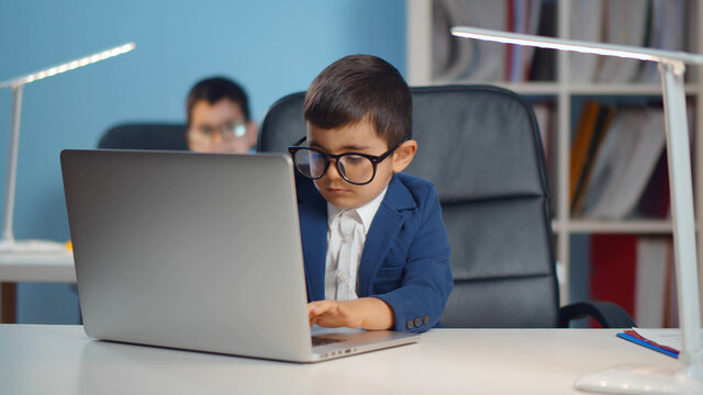 Adorable preschool child working on laptop at table in suit and glasses