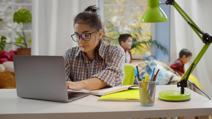 Young tired woman trying to work on laptop at home office with kids playing on background