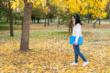 young woman with curly black hair wears blue wireless headphones and in hand her laptop to work outdoors. park in autumn with dry leaves fallen from the trees