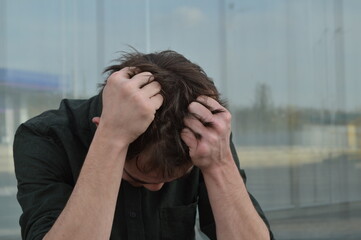 A young man in a khaki shirt grabbed his hair with both hands against the background of a glass wall. Despair
