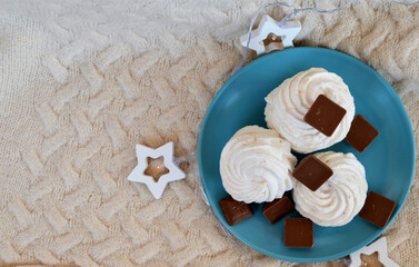 Handmade marshmallows and pieces of milk chocolate on a pale turquoise plate, white wooden stars on a cream-colored knitted sweater background. View from above. New Year. Winter. Christmas
