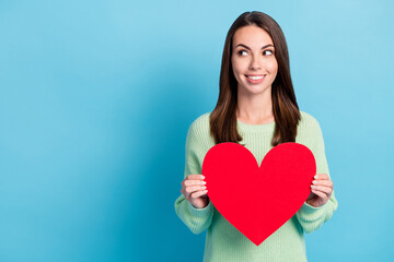 Photo portrait of curious pretty girl holding red symbol of love big heart wearing green jumper smiling looking up isolated on vibrant blue color background with empty space
