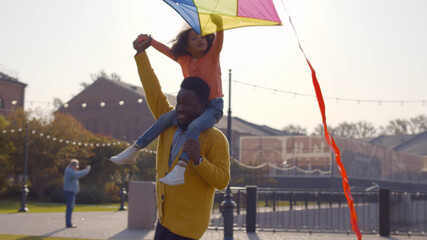 Cute african little girl sitting on shoulders of dad and playing with colorful kite