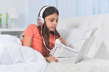 Beautiful young girl in headphones on soft beige bed with laptop