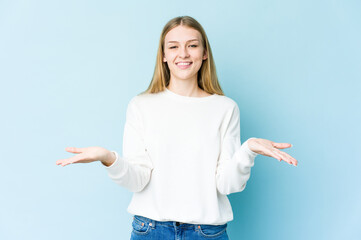 Young blonde woman isolated on blue background holding something with palms, offering to camera.