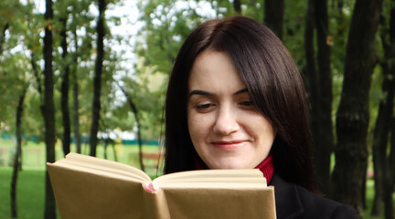 Fototapeta premium Smiling brunette woman reading a book in the park. Portrait of a happy charming Caucasian woman outdoors.