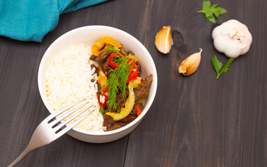 Roast beef with sweet pepper and white rice. White plate, garlic in the background, dark background.