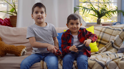 Cute preschool boys playing video game holding joysticks sitting on couch at home