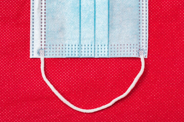 Fragment of blue medical face mask on background of non-woven fabric bright red color. Flat lay, close-up view of concept problem lockdown, coronavirus pandemic covid-19 disease, medicine quarantine.