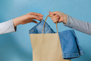 One hand of the child is passing a craft paper bag with blue jeans to the other hand. Concept of thrift stores, resale, second hand. - obrazy, fototapety, plakaty