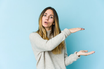 Young caucasian woman isolated on blue background shocked and amazed holding a copy space between hands.