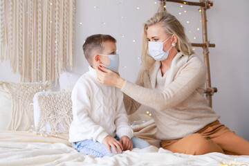 Obraz na płótnie Canvas A beautiful mother and son in medical masks are having fun at home near a Christmas tree in a white interior. Family happiness, holiday, joy, vacation, games with a woman. New Year's preparations.