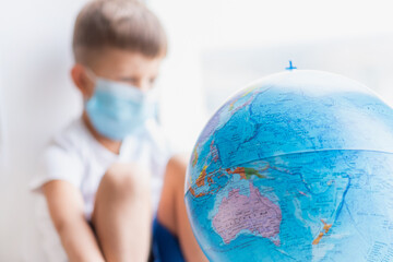 The masked kid sits at home in quarantine. The child is examining the globe while sitting on the windowsill. Prevention of coronavirus Covid-19 during a pandemic.