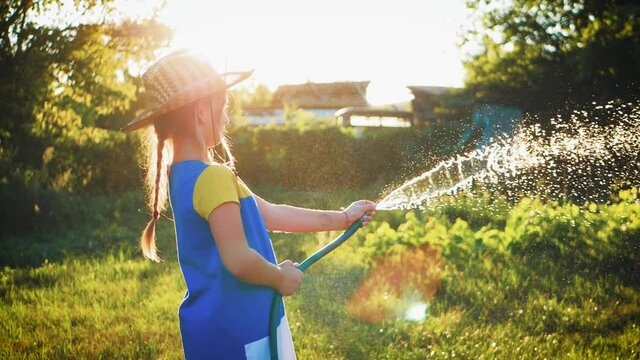 Funny little girl in hat playing with garden hose in sunny backyard. Adorable little girl playing with a garden hose on hot and sunny summer evening. Summer outdoors activity for kids.