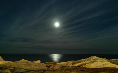 Fototapeta na wymiar bsh Luna tra le nuvole sul mare di sera vista dagli scogli con riflessi di luce sull’acqua in inverno