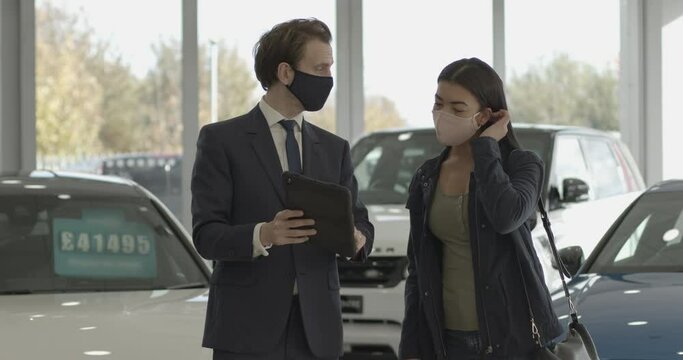Car sales person talking to customer wearing protective face mask in car dealership showroom and looking at digital tablet