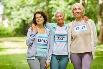 Group of three joyful senior girlfriends taking part in summer marathon race standing together in park smiling at camera