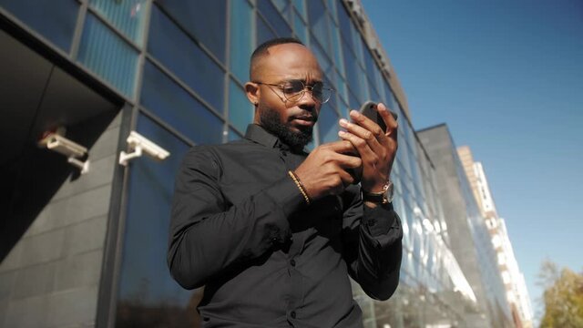 Millennial african man holding smartphone, swiping photos in online dating app. Young black man ordering taxi in mobile application, web surfing typing message in social networks.