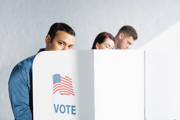 indian man looking at camera from polling booth near multicultural electors on blurred background