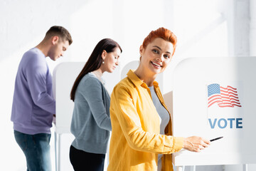smiling woman pointing with pen on american flag and vote inscription on polling booth on blurred...
