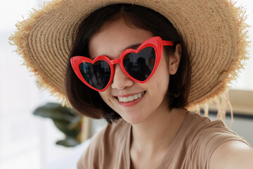 short haired Asian woman wearing a brown T-shirt in a straw hat and red heart-shaped sunglasses is taking selfie photo of him in the bedroom with a smartphone. concept of travel in summer and holiday.