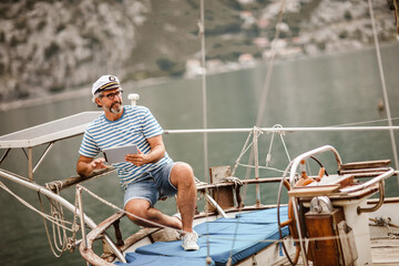 Mature man standing at helm of sailboat out at sea using digital tablet on a sunny afternoon.