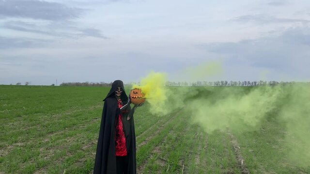 Woman stands in Halloween costume of death and holds jack-o-lantern with yellow smoke from it.