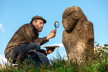 Scientist historian examines  stone sculpture on  mound through magnifying glass