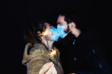 Young couple wearing protective face masks for Coronavirus and holding sparklers for New year's...