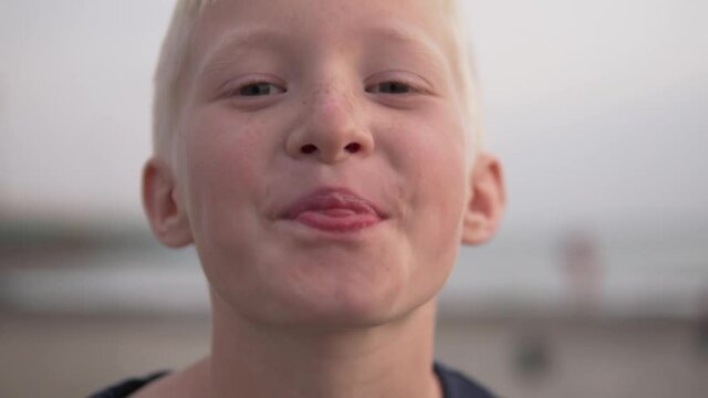A close-up of a beautiful albino boy shows his tongue against a backdrop of a blurred sea.
