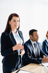 Smiling female politician gesturing, while standing in boardroom with blurred colleagues on...