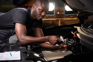 african professional auto service technician in uniform standing near car hood repairing and using...