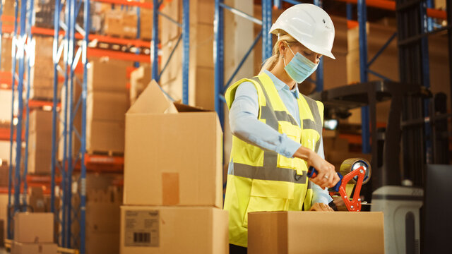 In Retail Warehouse Professional Worker Wearing Facial Mask Packing Parcel, Cardboard Box Sealed With Tape Dispenser Ready For Shipment. Delivery And Distribution Center Full Of Shelves With Products