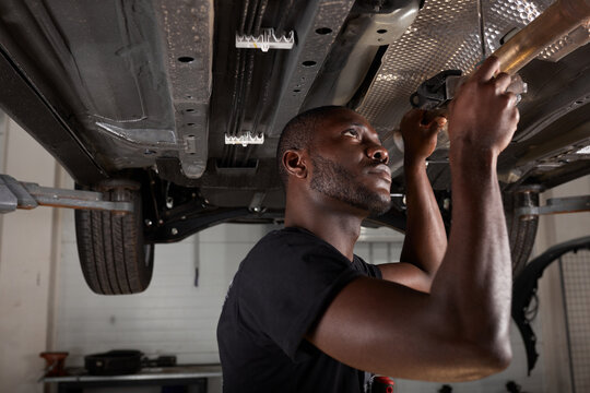 Young Male Car Mechanic In Uniform Checking Car In Automobile Service With Lifted Vehicle, Handsome Hardworking Guy Working Under Car Condition On Lifter. Automotive Car Repair Concept