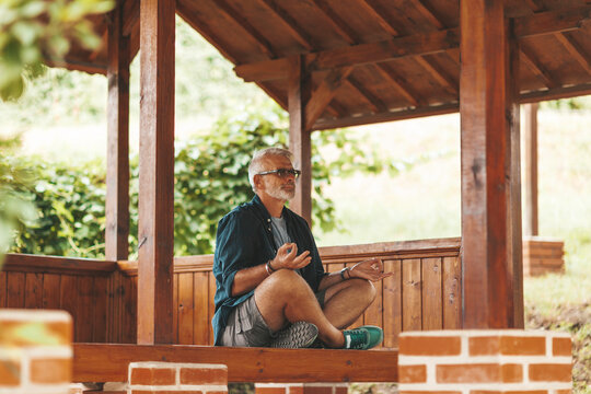 A 50-year-old Bearded Man Relaxes In The Lotus Position On The Terrace Of A House. Rest From Worries, Peace Of Mind In Retirement.