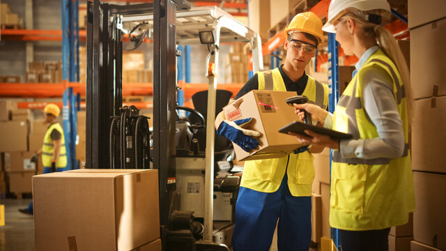 In Warehouse Manager Uses Digital Tablet and Scans Cardboard Boxes for Inventory, Talks with Forklift Driver about Package Delivery. Workers in Global Distribution Center with Shelves with Goods 