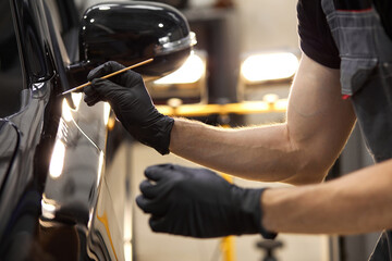 cropped caucasian auto service worker is painting car details, use small brush and paint, apply it on surface of black automobile