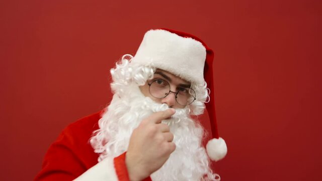 Funny Santa taps his finger on the camera and points aside at the copy space. Santa at Christmas asks to look at the recommendation on the background of the red wall.