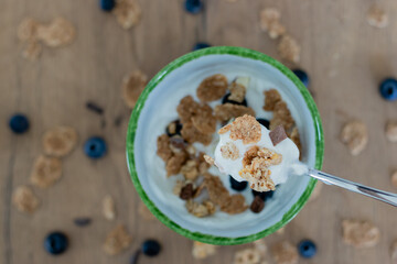 Healthy breakfast with greek yogurt, cereal and blueberries in a spoon with bowl and scattered blueberries and cereal on a counter top underneath. 