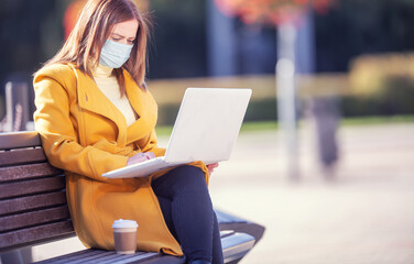 Woman sits on a bench outdoors working on a laptop in face mask on a sunny fall day