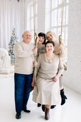 Christmas, winter holidays, family togetherness. Smiling middle aged mom sitting on swing, dad and two young pretty sisters, having fun while celebrating holidays at beautiful light decorated hall.