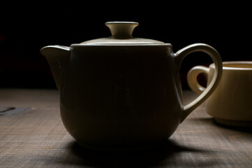 Still life, chiaroscuro renaissance, or baroque style silhouette of a teapot, tea pot and a tea cup on a wooden vintage table and a black background, abstract image 